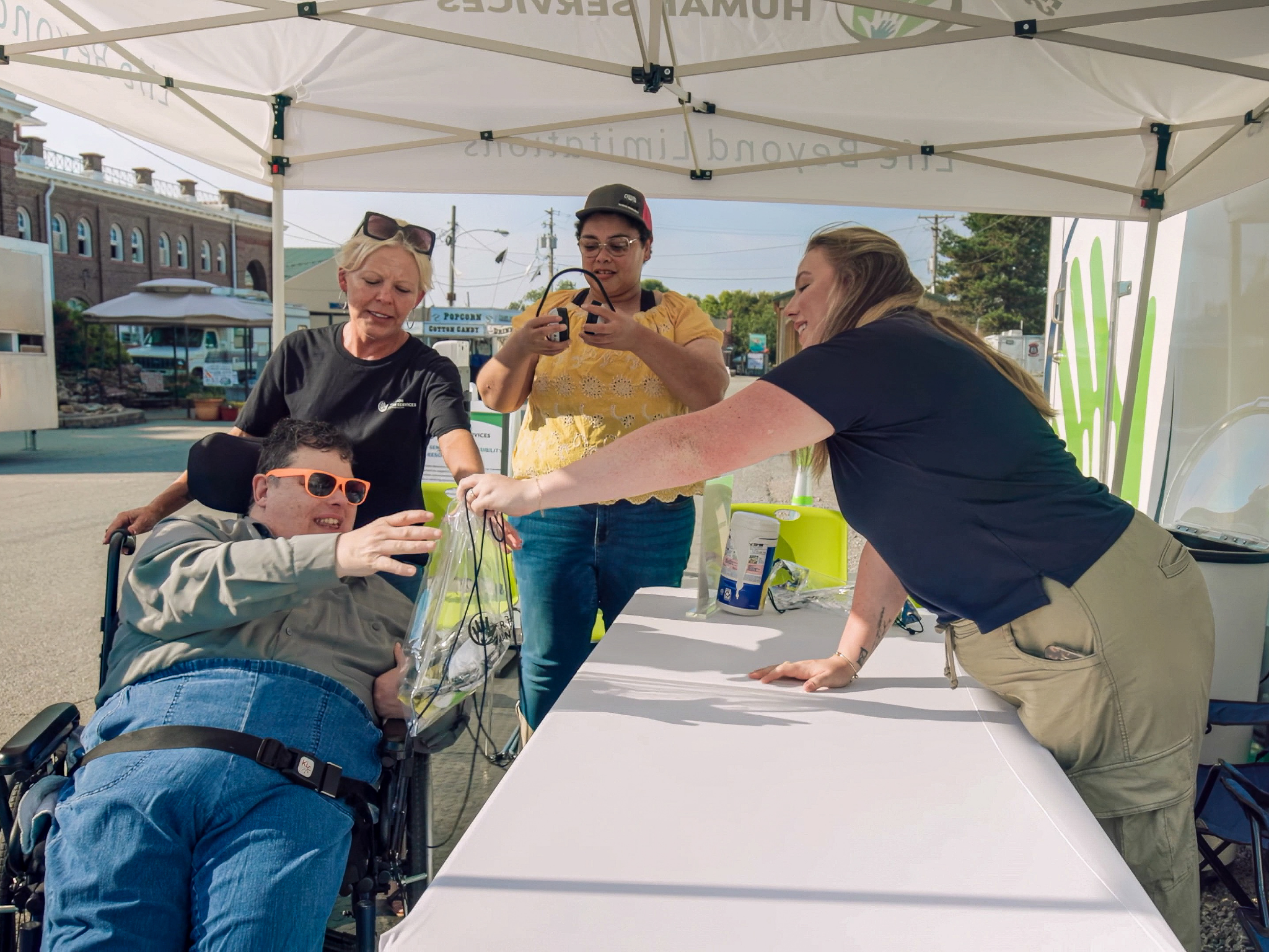 Staff assist a man in a wheelchair reach items at an outdoor resource table.