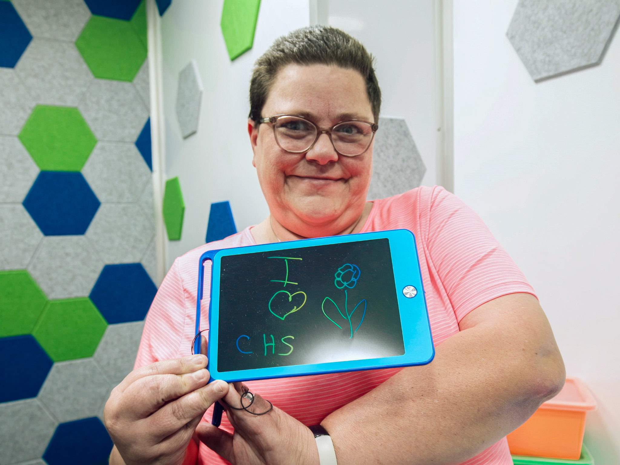 A woman holds a tablet displaying the message ‘I heart CHS’ with a simple flower drawing.