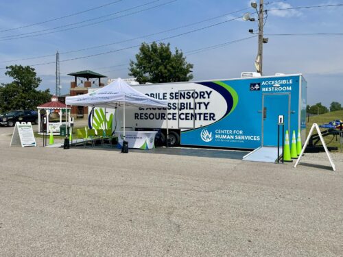 A large blue mobile trailer labeled “Mobile Sensory Accessibility Resource Center” is parked in an open lot. The trailer has a wheelchair-accessible restroom with a ramp and signage. In front of the trailer, there is a white canopy tent with the “Center for Human Services” logo and the slogan “Life Beyond Limitations.” Under the tent are bright green chairs and an information table covered with a branded tablecloth. Green traffic cones and sandwich board signs provide additional information for visitors. The setup is designed to provide community outreach and accessible services in a mobile format.