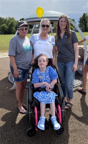 Four people gathered outdoors at a playground event, with a child in a wheelchair at the center, surrounded by supportive adults.