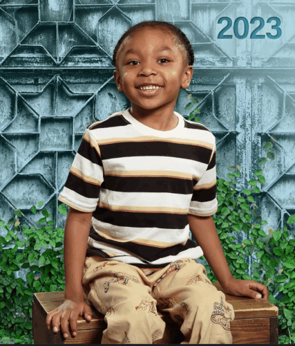 A boy sits smiling for his school photo in a black and white stripped shirt and khaki pants.