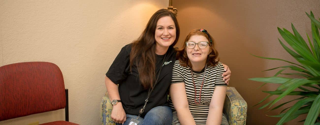 Untitled design A service coordinator with long brown hair sits beside a young woman with red hair and glasses, smiling together in a warmly lit room.