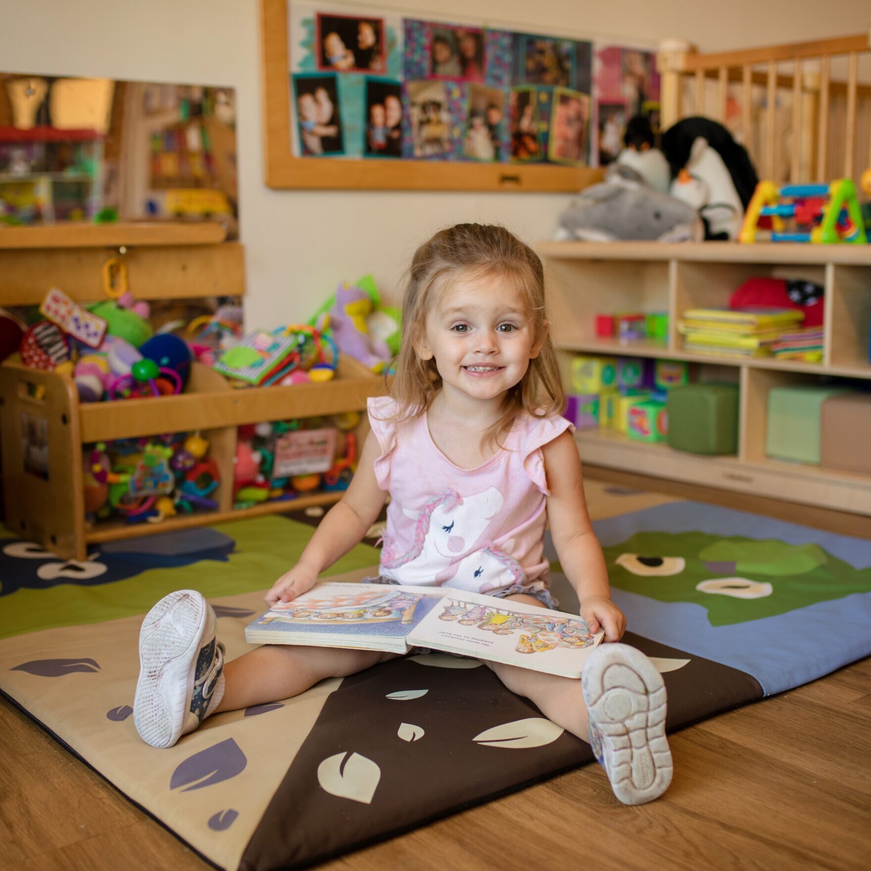 A little girl in a pink dress sits on the floor smiling with a book in her lap
