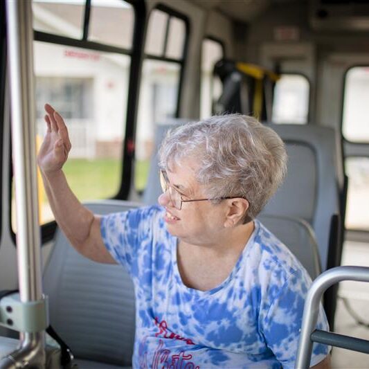 An older lady with glasses waves out the window while sitting on a bus.