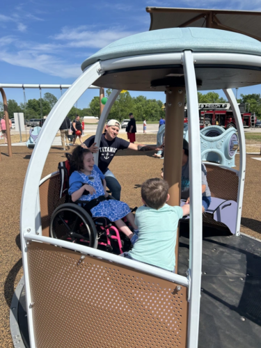 A group of children, including a girl in a wheelchair, play together on an inclusive playground spinner under the supervision of an adult. The atmosphere is joyful and sunny, with a modern, accessible playground in the background and families enjoying the day.