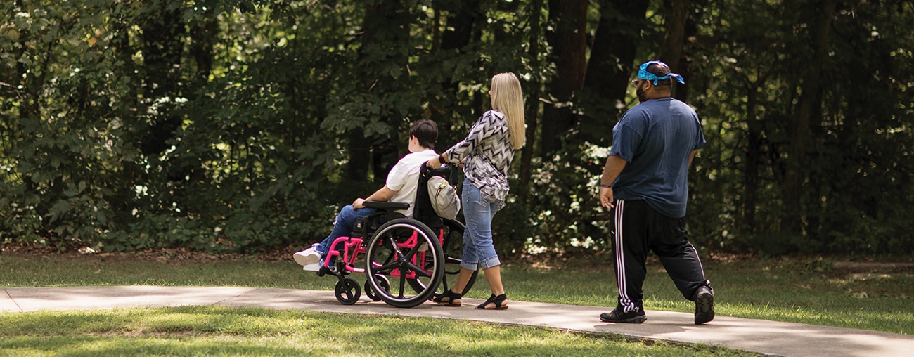 A woman pushing a person in a wheelchair on a walk outside