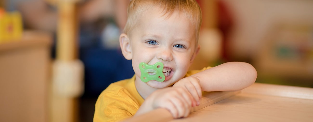 A smiling young boy with a pacifier in his mouth.
