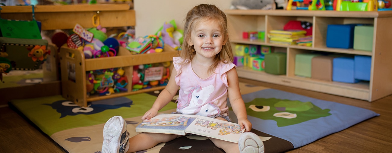 A smiling young girl reading a book in her lap.